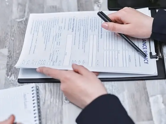 Close-up of hands reviewing an insurance policy document on a clipboard, with a pen and notepad beside a laptop