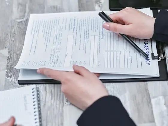Close-up of hands reviewing an insurance policy document on a clipboard, with a pen and notepad beside a laptop
