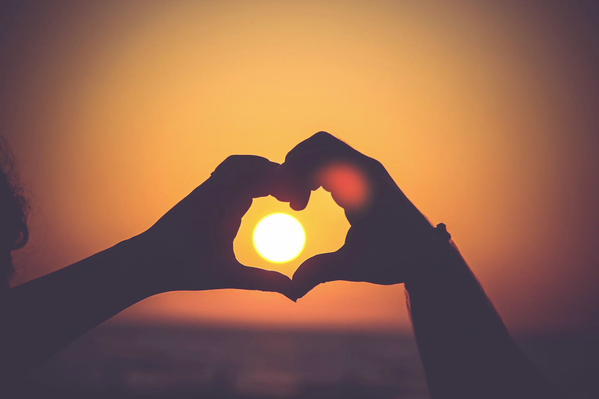 A couple making a love heart with their hands with a sunset in the background.